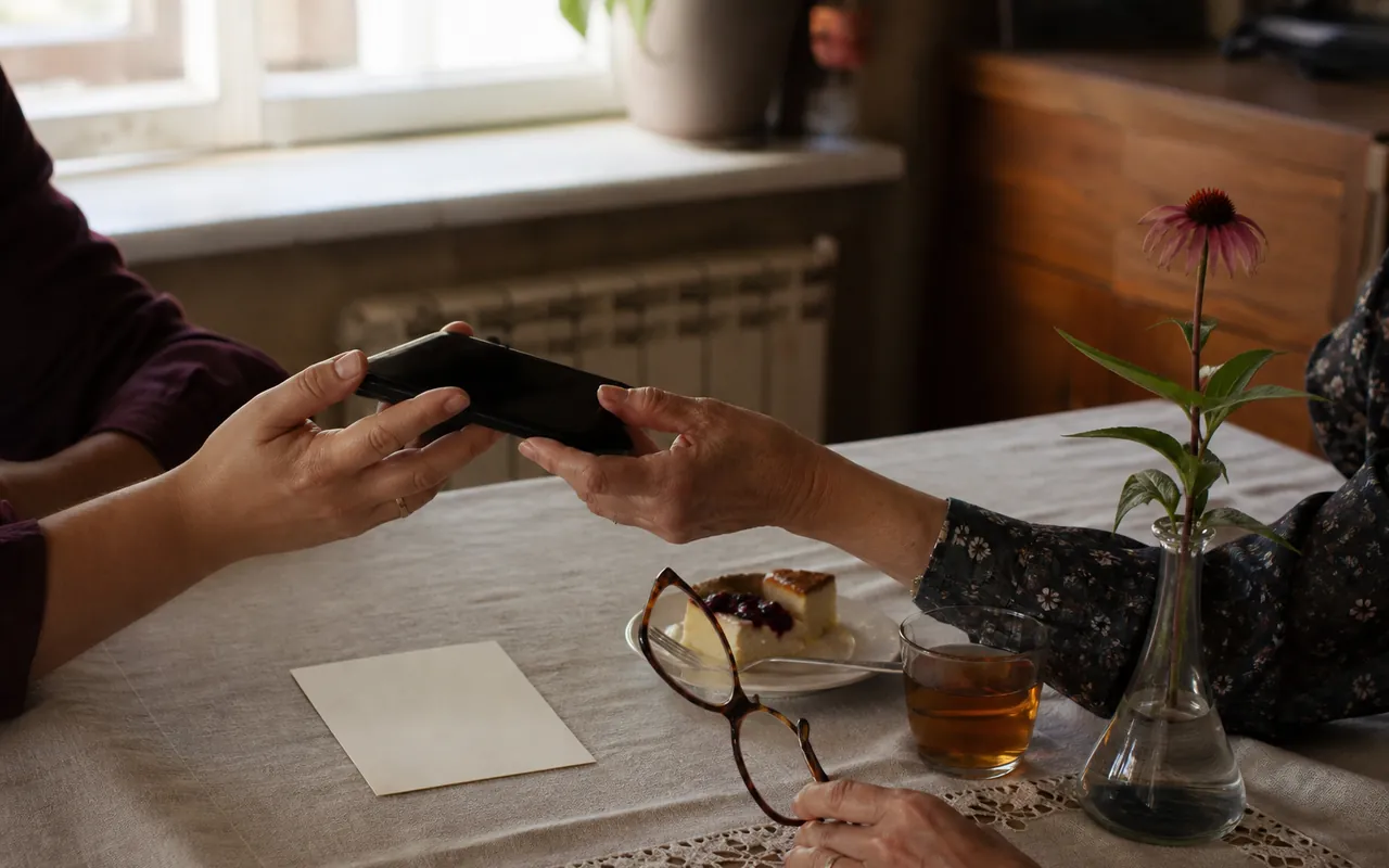 Dwie osoby przekazują sobie telefon nad stołem, obok leży papierowa karta