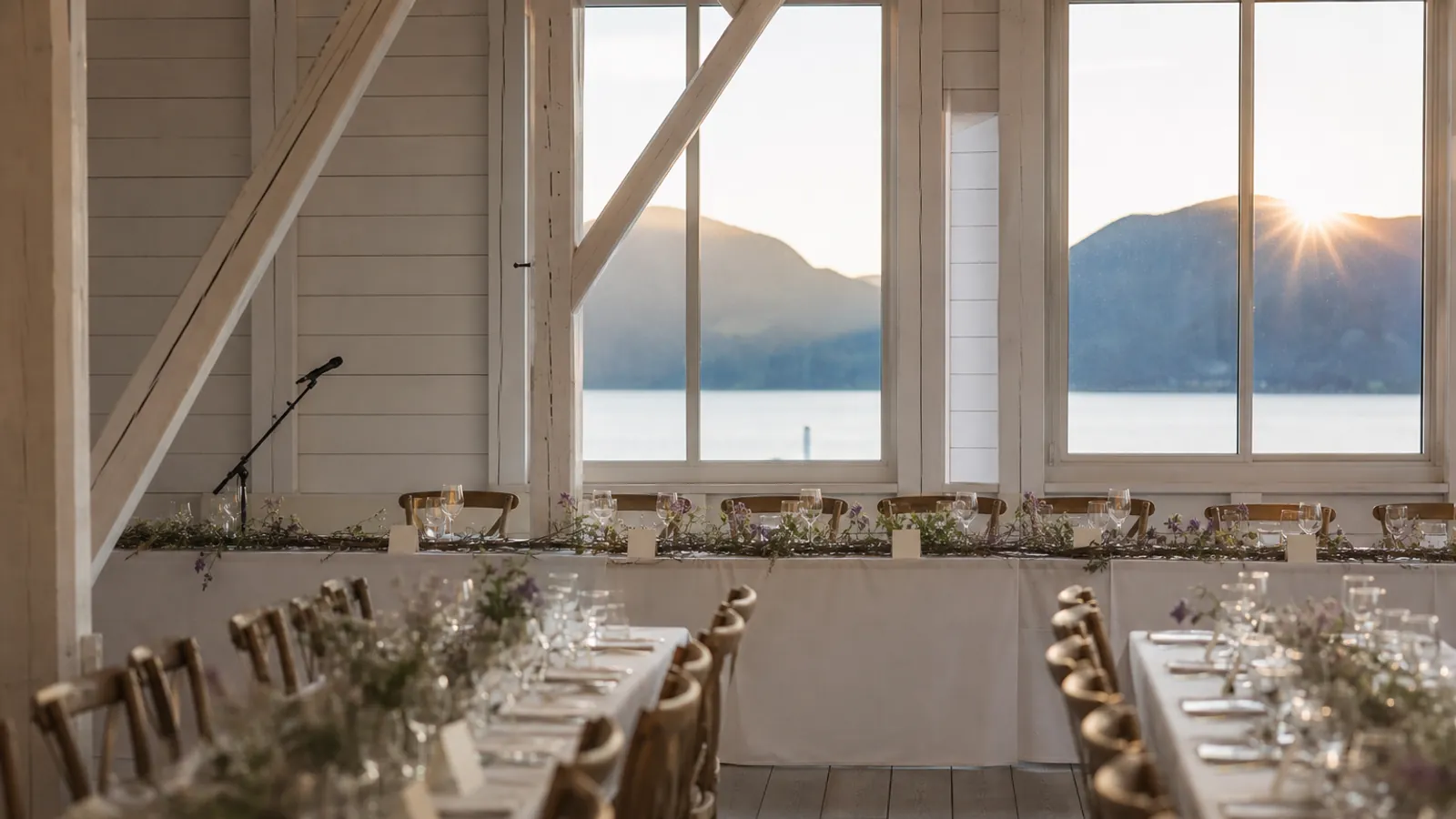 Et langt hovedbord dekket på én side i en hvitmalt låve, med bordkort, lave blomster og kveldslys fra fjorden utenfor